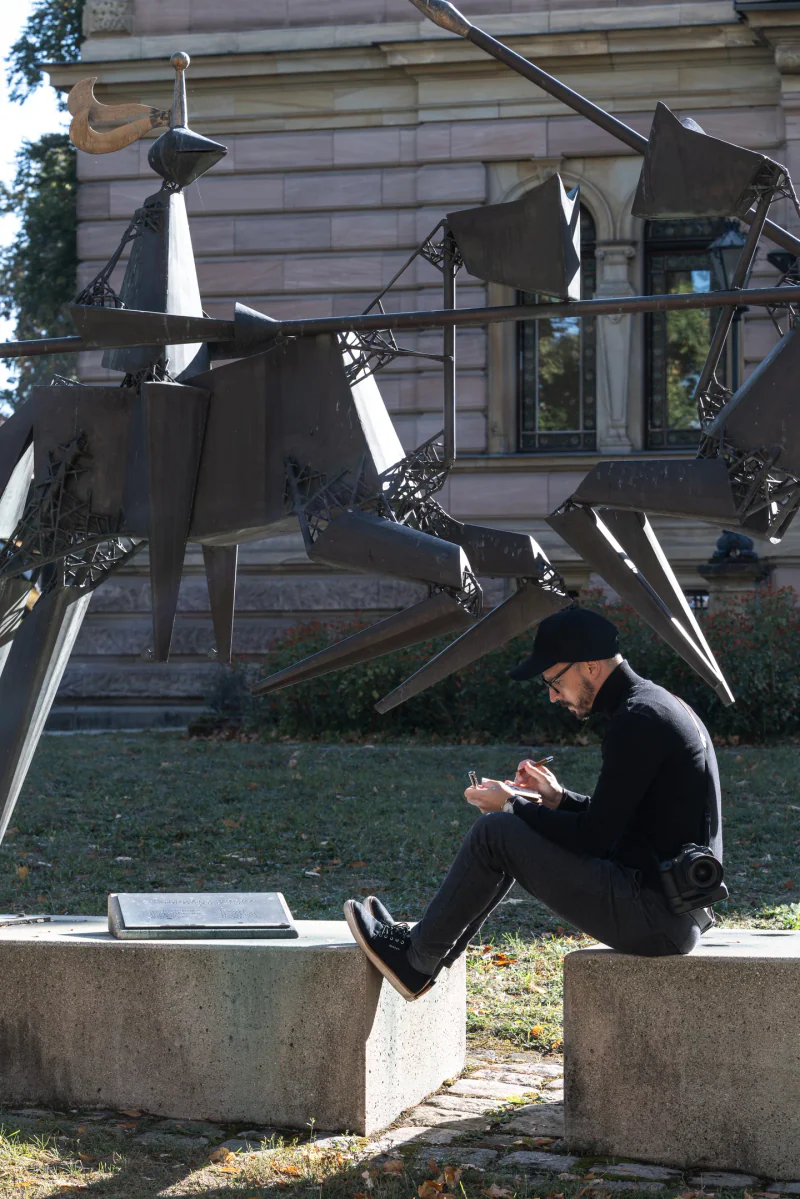 Filip writing in his notebook beneath the large jousting-knights sculpture.