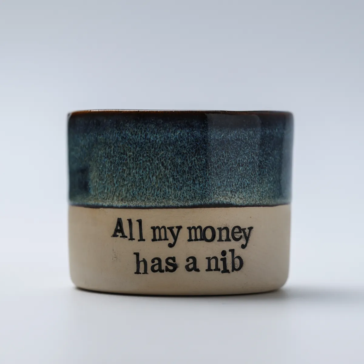 Front view of All My Money Has a Nib mug with textured blue glaze and black lettering
