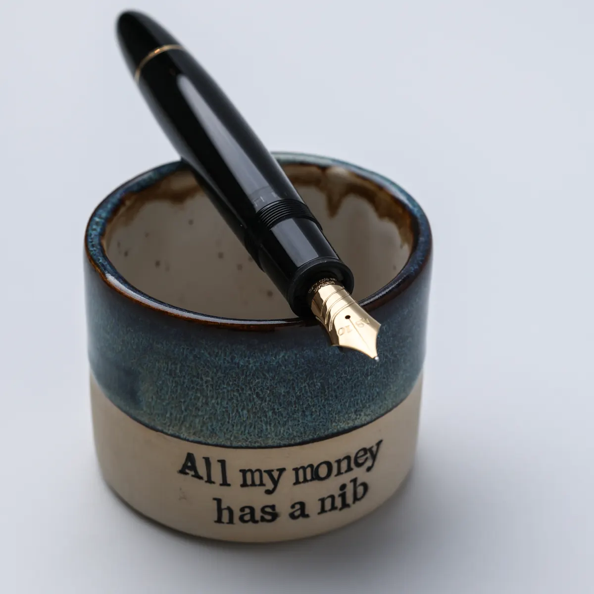 Black fountain pen with a gold nib resting across the rim of an All My Money Has a Nib mug