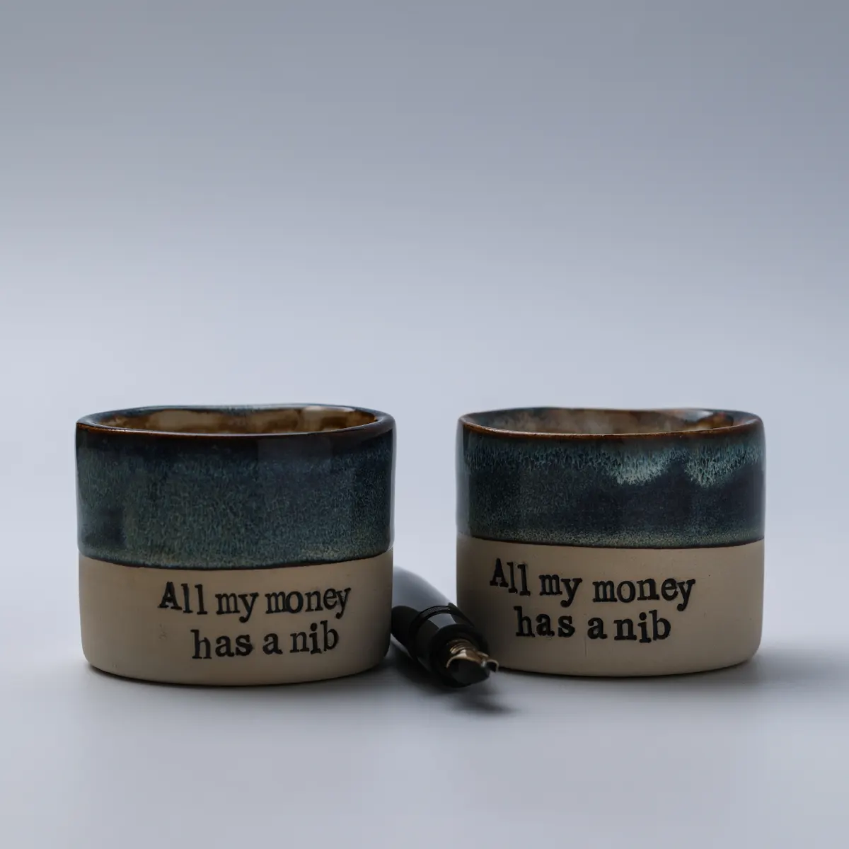 Two All My Money Has a Nib ceramic mugs side by side with an uncapped fountain pen between them
