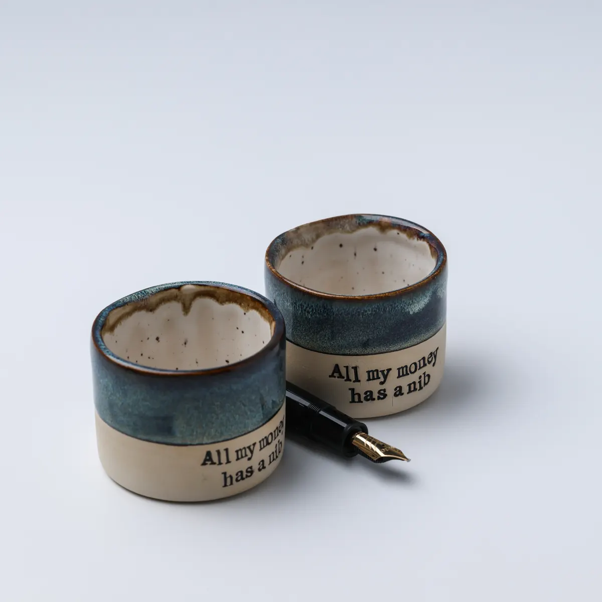 Elevated view of two All My Money Has a Nib mugs and a fountain pen, showing the speckled interiors