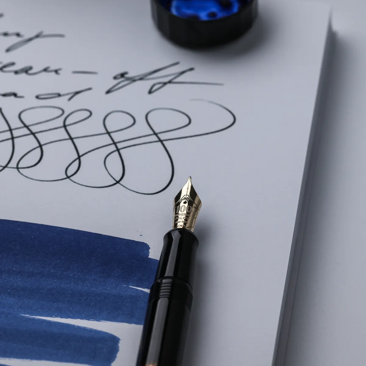 Close-up of a gold Pilot fountain pen nib beside handwritten loops and a blue ink swatch on FMP paper