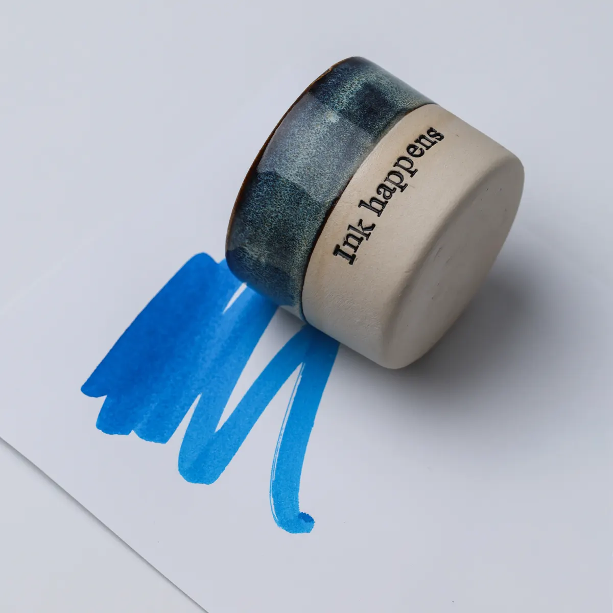 Ink Happens mug resting on its side on paper with blue ink swatches