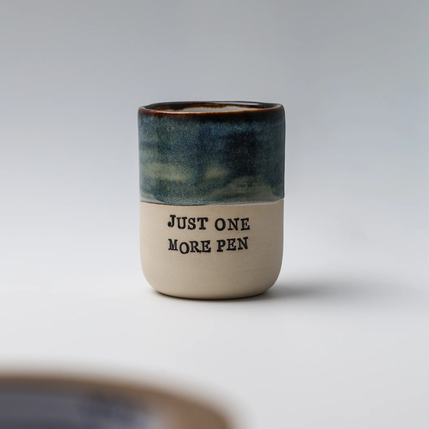Front view of an empty Just One More Pen ceramic cup showing textured blue glaze and black lettering on natural clay