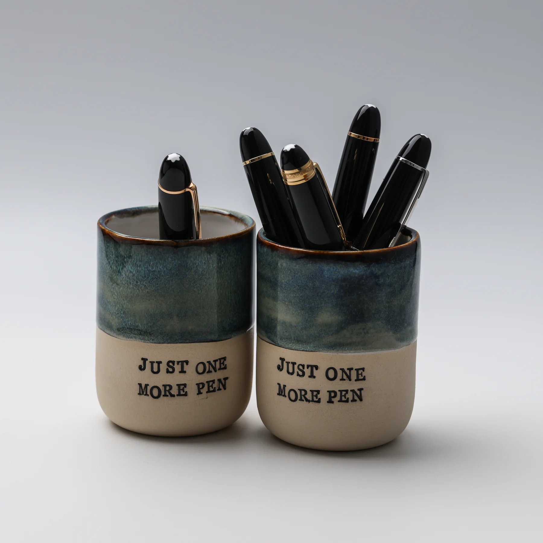 Two handmade Just One More Pen ceramic cups with blue glaze and natural clay bases, holding fountain pens